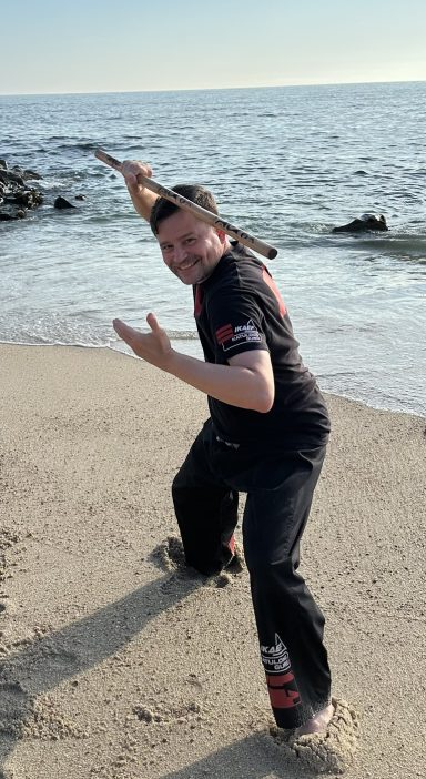 Mann in Strandposition mit einem Stock, Wasser im Hintergrund.