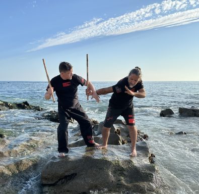 Zwei Personen in Schwarz, die mit Stöcken auf Felsen am Wasser kämpfen.
