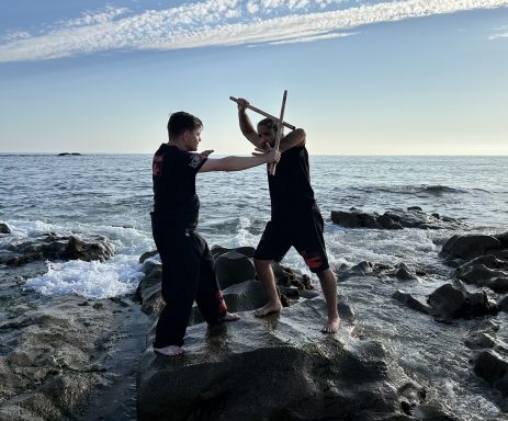 Zwei Personen kämpfen mit Stöcken auf Felsen am Meer bei Sonnenlicht.